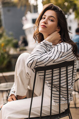 Cute young caucasian girl posing looking at camera relaxing outdoors in outdoor cafe. Brunette woman with wavy hair wears jacket and pants in spring. Leisure lifestyle and beauty concept.