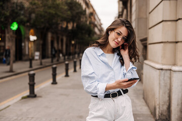Fototapeta premium Nice caucasian young woman is using headphones and smartphone standing on street. Brunette with wavy hair wears casual clothes. Telephone technology concept