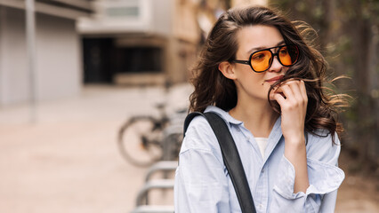 Calm young caucasian woman looking away relaxing outdoors in spring. Brunette girl with wavy hair wears sunglasses, casual clothes. Leisure lifestyle and beauty concept.