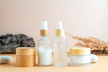Fototapeta na wymiar White frosted glass cosmetics bottles and jars with pamboo lids with tree bark, pampas grass, stones on beige background. Autumn fall beauty products set.