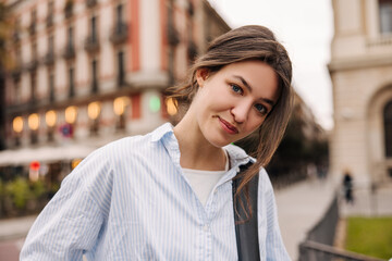 Beautiful young european lady looking at camera spending leisure time outdoors on vacation. Brunette wears casual blue shirt. City life concept