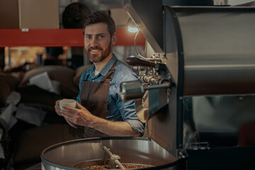 Smiling business owner with cup of fresh coffee sitting near roasting machine on own factory