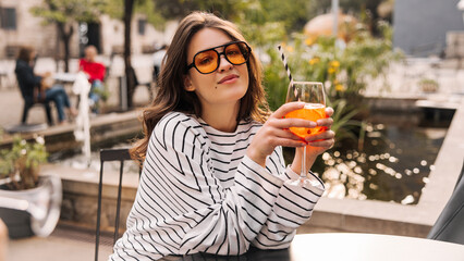Pretty caucasian young lady is enjoying cooling drink sitting in cafe on hot day. Brunette wears sunglasses, light clothes. Relaxation concept