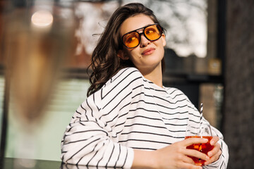 Beautiful young caucasian girl spends leisure time outdoors with cooling drink. Brunette with wavy hair wears sunglasses, white sweatshirt. Lifestyle concept