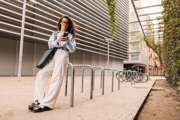 Full length focused young caucasian woman using smartphone standing outdoors. Brunette with wavy hair wears classic clothes. Technology concept
