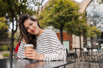 Cute young caucasian woman looks at camera, spends leisure time in cafe with cup of coffee. Brunette with wavy hair is relaxing outdoors. Lifestyle concept