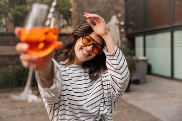 Happy young caucasian girl holds out glass to camera posing outdoors. Brunette wears sunglasses, white blouse. Summer playful mood concept