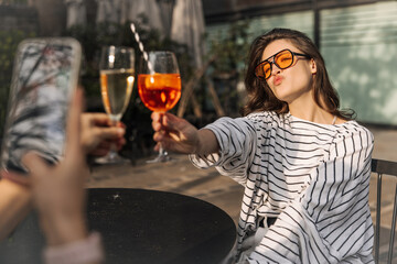Cute caucasian young woman pursing her lips into kiss raises glass sitting in outdoor cafe. Brunette wears sunglasses, white blouse. Summer vacation concept.