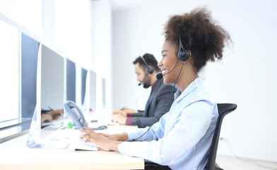 Fototapeta premium Happy attractive business people. Call center worker accompanied by team. Smiling customer support operator at work. Young employee working with a headset. Successful business professionals.