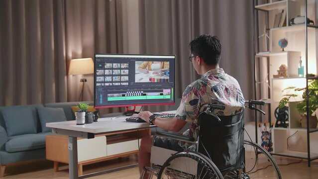 Back View Of Asian Man In Wheelchair Is Using A Desktop Next To The Camera Editing The Video At Home

