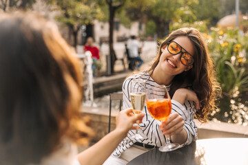 Nice european young lady drinking non-alcoholic cocktail relaxing sitting in street cafe. Brunette girl with wavy hair wears sunglasses, casual clothes. City life concept