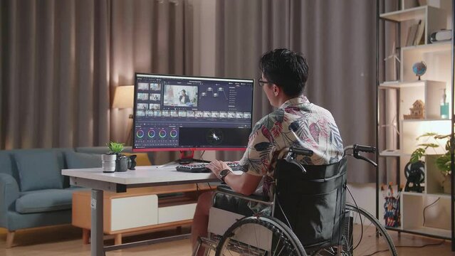 Back View Of Asian Man In Wheelchair Shaking His Head And Having A Headache While Editing And Color Grading The Video By A Desktop Next To The Camera At Home
