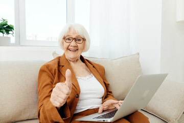 a happy, emotional elderly woman with gray hair is sitting in a bright apartment on a beige cozy sofa near the window happily working at a laptop showing a thumbs up