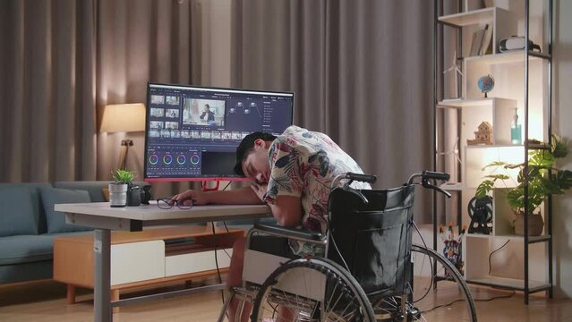 Back View Of Asian Man In Wheelchair Sleeping While Editing And Color Grading The Video By A Desktop Next To The Camera At Home
