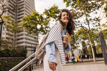 Happy young caucasian woman with coffee, notebooks and backpack goes to class on street. Brunette in good mood wears shirt, sweatshirt and trousers. People emotions, lifestyle and fashion concept.