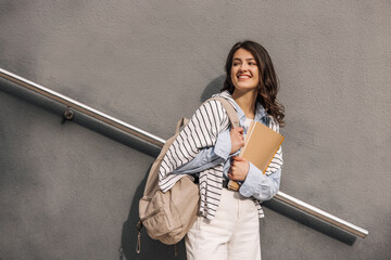 Stylish young caucasian female student with notebook and backpack looking away on grey background. Brunette woman with wavy hair wears classic clothes. Lifestyle concept