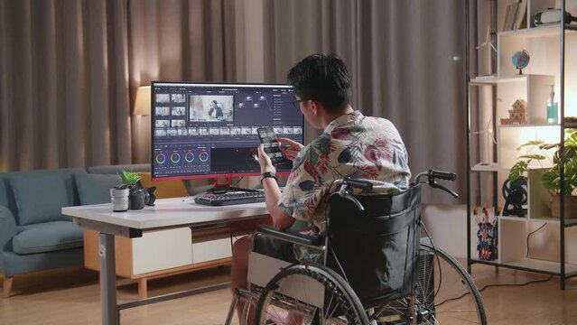 Back View Of Asian Man In Wheelchair Using Smartphone While Editing And Color Grading The Video By A Desktop Next To The Camera At Home
