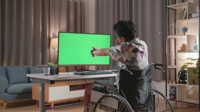 Back View Of Asian Man In Wheelchair Stretching While Using Mock Up Green Screen Desktop Next To The Camera At Home
