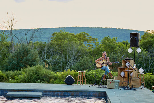 Candid image of a young woman playing acoustic guitar with trees and mountains in the background and a pool in the foreground. 