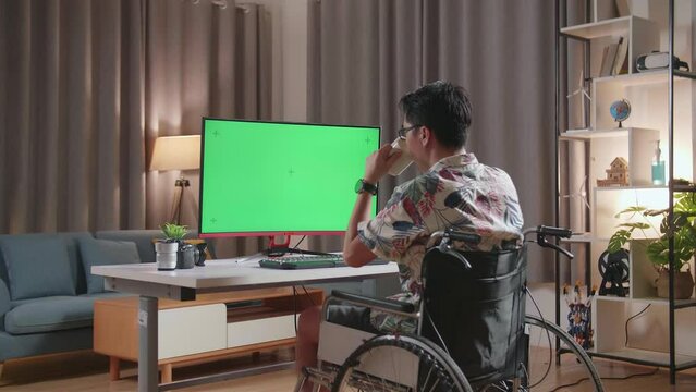 Back View Of Asian Man In Wheelchair Drinking Coffee While Using Mock Up Green Screen Desktop Next To The Camera At Home
