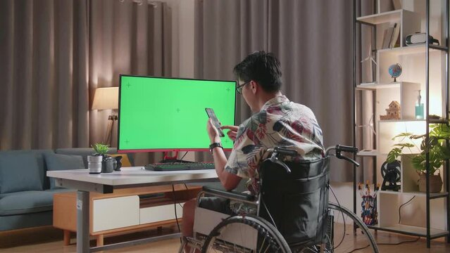 Back View Of Asian Man In Wheelchair Using Smartphone While Using Mock Up Green Screen Desktop Next To The Camera At Home
