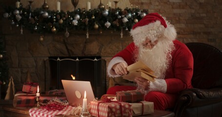 Santa claus in a red suit reads letters to children by the fireplace in his residence