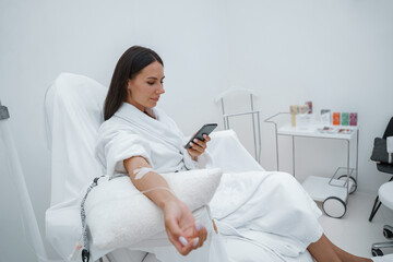 Beautiful woman in white bathrobe use phone during medical procedure in beauty clinic - obrazy, fototapety, plakaty
