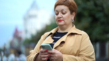 An elegant woman with short red hair and a raincoat is standing on the street and using a smartphone - 532854104