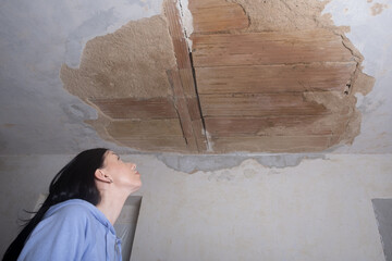 a young woman looking up at a ruined hole in the ceiling. The plaster fell off from moisture. Old apartment  restoration repair and fixing. - obrazy, fototapety, plakaty