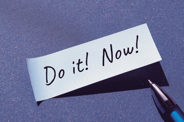 Do it now motivator message written on sticky paper, office sticker with pen. Single simple minimal motivation note stiicker. Handwriting on blue paper background.
