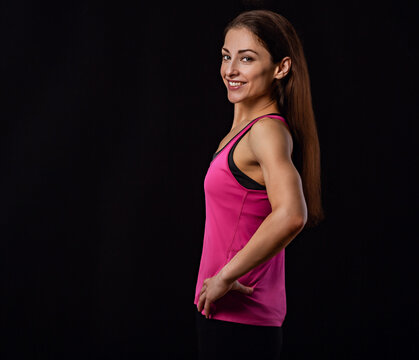 Happy sportswoman in pink sportsbra and shirt, holding hands on waist, fitness trainer with long healthy hair standing in power pose, workout in gym isolated on black background