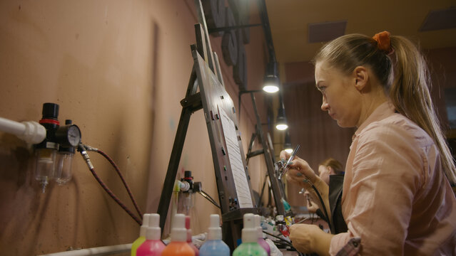 Woman artist learns to paint with airbrush with acrylic dye, paper and easel. Indoors. Concept modern art, airbrushing, aerography, draw spraying picture create sprayer drawing graffiti aerosol spray
