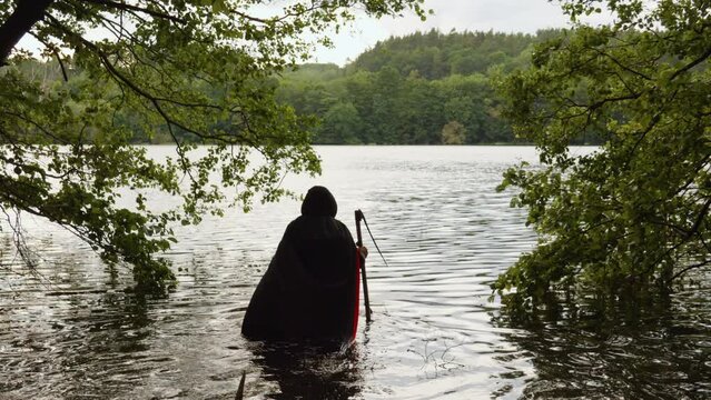 Back View Of Grim Reaper In Black Hooded Robe Walking Into The Water With Scythe. wide