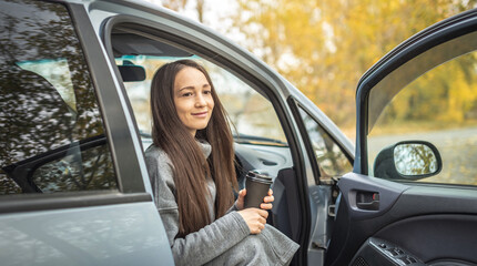 Woman is sitting in a car with a cup of coffee and bouquet of yellow autumn leaves, admiring the autumn landscape and enjoying the silence and beauty of nature