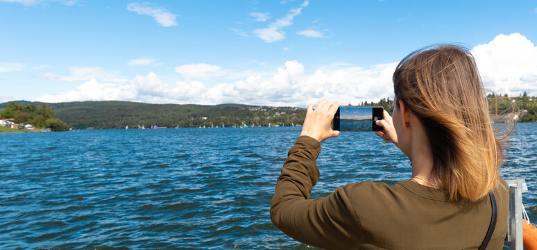 Photo of a young attractive woman taking pictures of a lake on a smart phone.