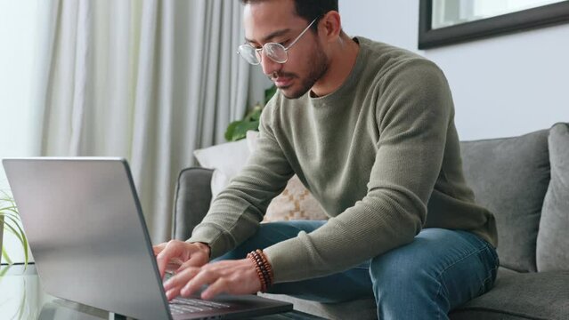 Man, glasses and laptop working on sofa in the living room typing email for business idea at home. Smart male freelancer or typist focused at work on computer sitting on couch planning design ideas