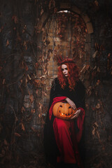 A red-haired young woman in a long black dress with a hood and gothic make-up holds a Halloween...