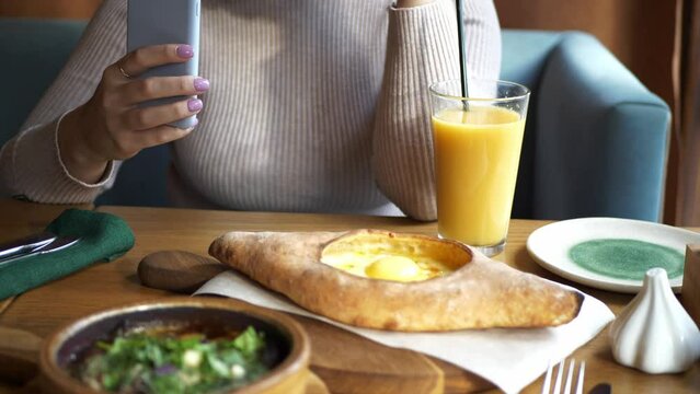 A girl in a cafe holds a freshly squeezed glass of orange juice and takes pictures of food on her smartphone. A young woman in a Georgian restaurant.