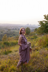 Beautiful young smiling girl in a long brown dress walks along the lawn. Happy woman walks at sunset on a hill overlooking the river. Concept of having rest in park during summer holidays or weekends