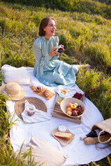 Laughing woman in a long dress with short hair sitting on a white blanket with fruits and pastries and looking at the pictures on camera. Concept of having picnic during summer holidays or weekends