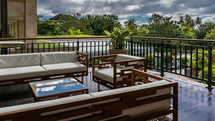 Fototapeta premium A seating area on the terrace. Wooden chairs and sofas with soft cushions are fenced with metal railings. The reflection of the sky on the glass of the coffee table. Green vegetation in the garden. 