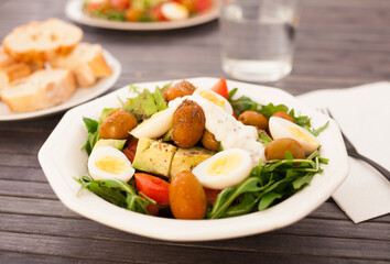 dish of fresh vegetables arugula, avocado, cherry tomatoes with olives and quail eggs on dining table