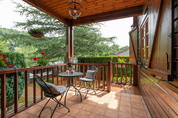 A wooden cottage with beautiful porch and table with two chairs. Cozy and nice house with many plants and trees on sunny day in european village.