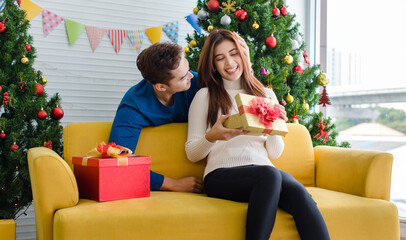 Asian boyfriend hugging cuddling girlfriend sitting smiling together holding present gift box on sofa in home living room full decorated with pine trees and flags celebrating Christmas eve festival