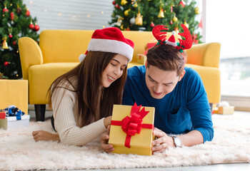 Obraz na płótnie Canvas Asian cheerful boyfriend wears deer antler headband and girlfriend in sweater and red Santa Claus hat laying down on floor looking each other eyes in decorated living room celebrating Christmas eve