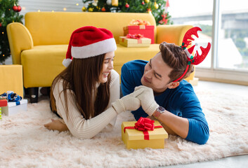 Asian cheerful boyfriend wears deer antler headband and girlfriend in sweater and red Santa Claus hat laying down on floor looking each other eyes in decorated living room celebrating Christmas eve