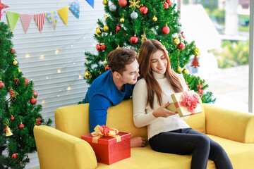 Obraz na płótnie Canvas Asian boyfriend hugging cuddling girlfriend sitting smiling together holding present gift box on sofa in home living room full decorated with pine trees and flags celebrating Christmas eve festival
