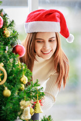 Asian happy cheerful female housewife in sweater and red Santa Claus hat standing smiling holding hanging shiny glitter golden ball decorating pine tree celebrating Christmas eve in home living room