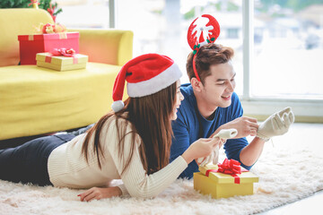 Asian cheerful boyfriend wears deer antler headband and girlfriend in sweater and red Santa Claus hat laying down on floor looking each other eyes in decorated living room celebrating Christmas eve