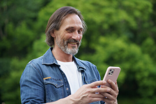 Happy attractive bearded man texting with his mobile phone in hands walking in park. Mature middle aged man swiping phone. Bearded man in denim t-shirt walking outdoor communicate by texting.